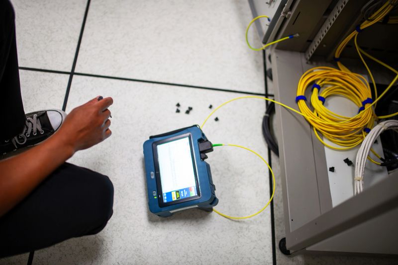 A technician kneels beside network equipment using a tablet for testing and diagnostics. Yellow cables surround the workspace, highlighting technology in action.