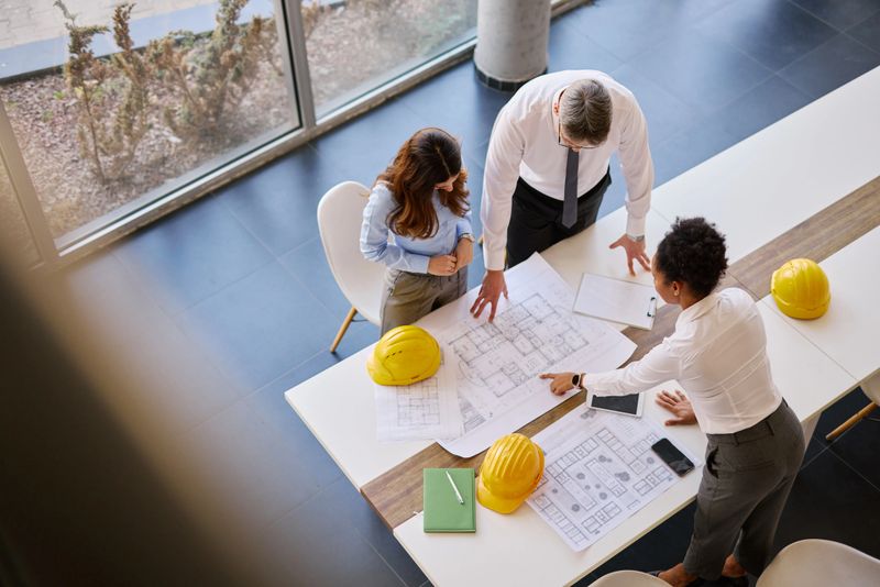 Three architects collaborating over blueprints on a table in a modern office, discussing strategies and planning details for an upcoming construction project