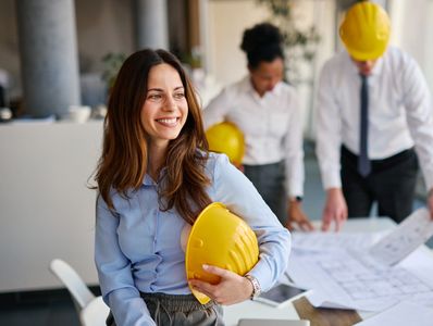 Smiling female engineer holding a yellow hard hat in an office.
