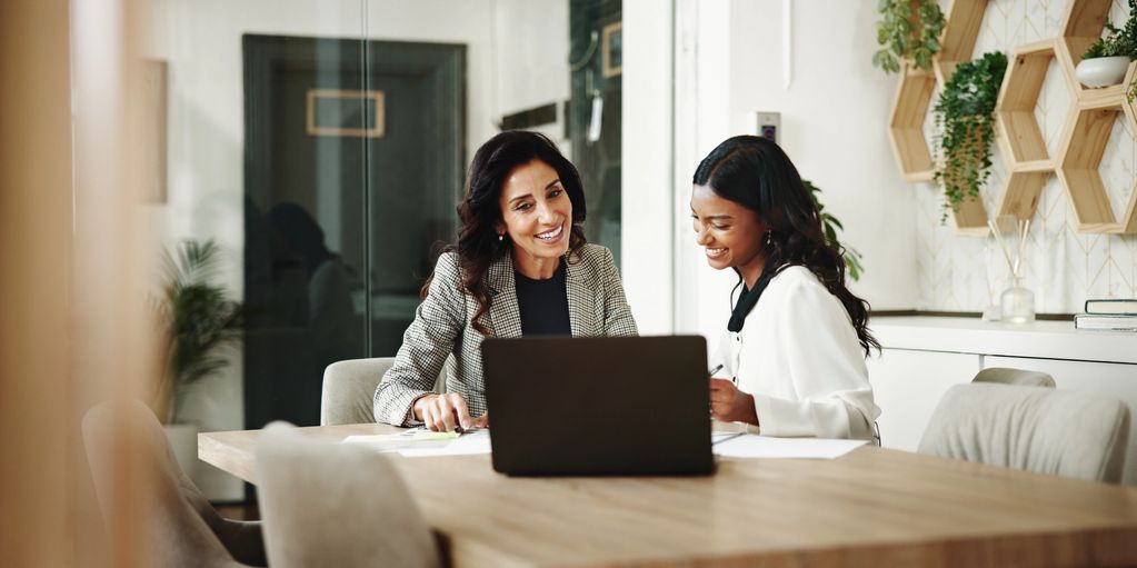 Two women smiling and discussing work at a laptop in a modern office.