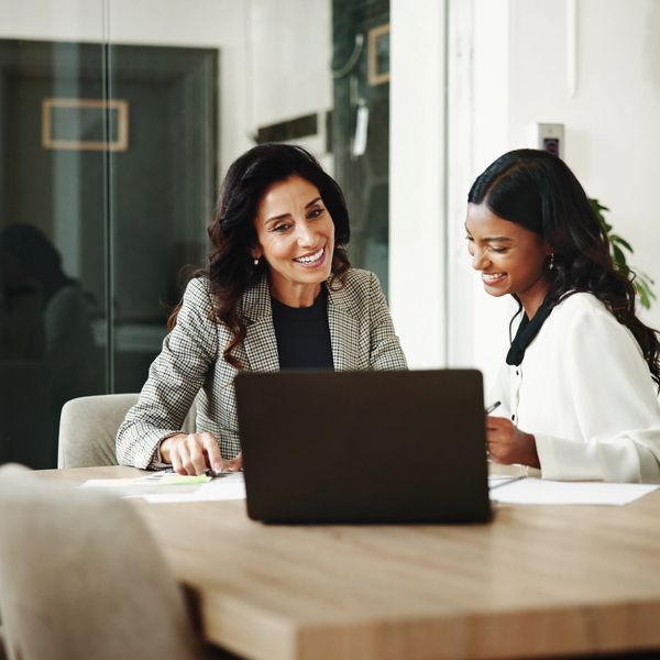 Two women smiling and discussing work at a laptop in a modern office.