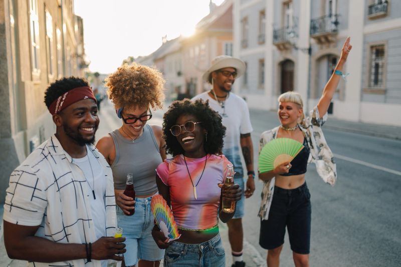 Group of cheerful multi ethnic friends enjoying a pride parade, holding rainbow hand fans and refreshing drinks while walking down a city street during a sunny day