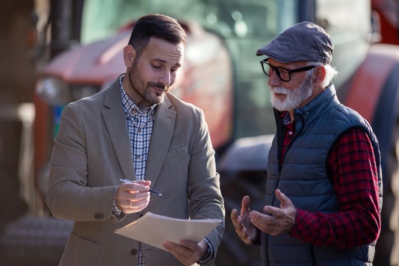 Business agent and senior farmer talking about loan contract with bank in front of tractor on ranch
