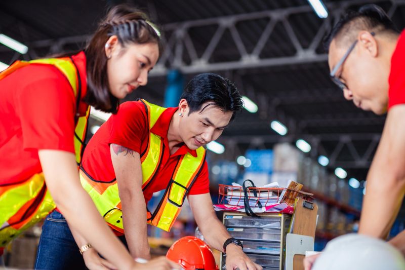 Three workers in red shirts and safety vests discuss plans while gathered around a table in a busy warehouse filled with boxes.