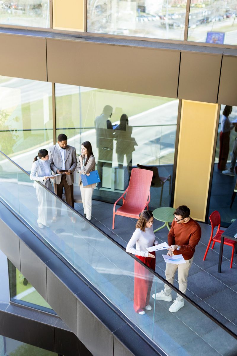 A group of professionals collaborates on a project while enjoying the airy space of a contemporary office balcony. Sunlight filters through large windows, enhancing their lively discussion.