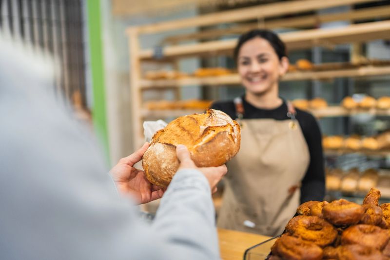 Baker handing a loaf of freshly baked bread to a customer in a bakery shop, with pastries in the foreground