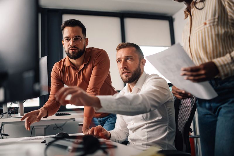Programmers cooperating on a project and analyzing code on computer, pointing at the screen while discussing the solution with a colleague holding a document