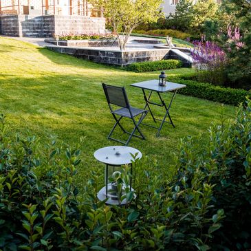 A cozy garden with a small table, chair, and lantern on lush green grass.