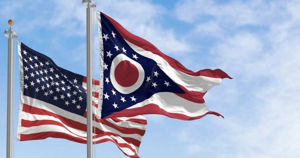 Ohio state flag and United States flag flying on poles against blue sky.