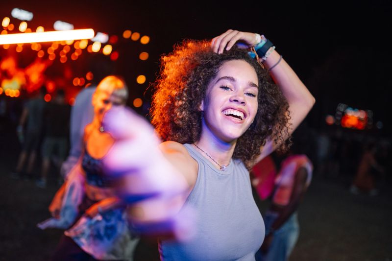 Happy young woman dancing joyfully at a music festival, reaching out with her hand to connect with friends, fully immersed in the vibrant atmosphere and electrifying live performance