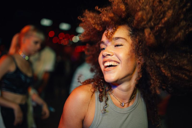 Young woman with curly hair is laughing and dancing with energy at a concert, surrounded by other people enjoying the music and nightlife