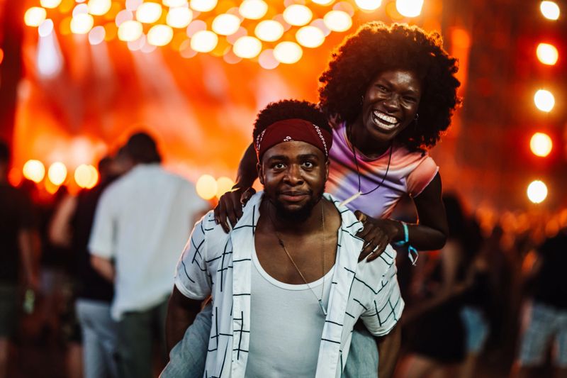 Two friends share a playful moment at a lively music festival, surrounded by colorful lights and a bustling crowd. The atmosphere is filled with excitement and laughter