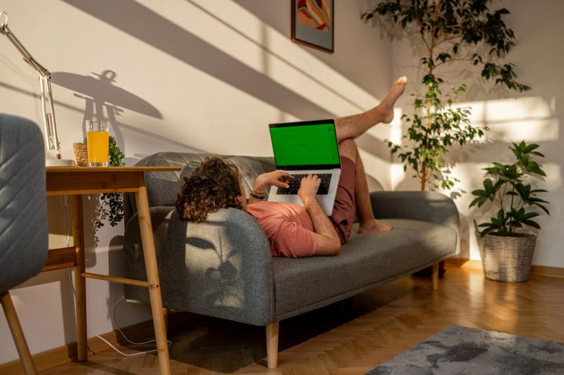 Young caucasian man lying on a sofa at home and using laptop , home leisure in sunset heat temperature