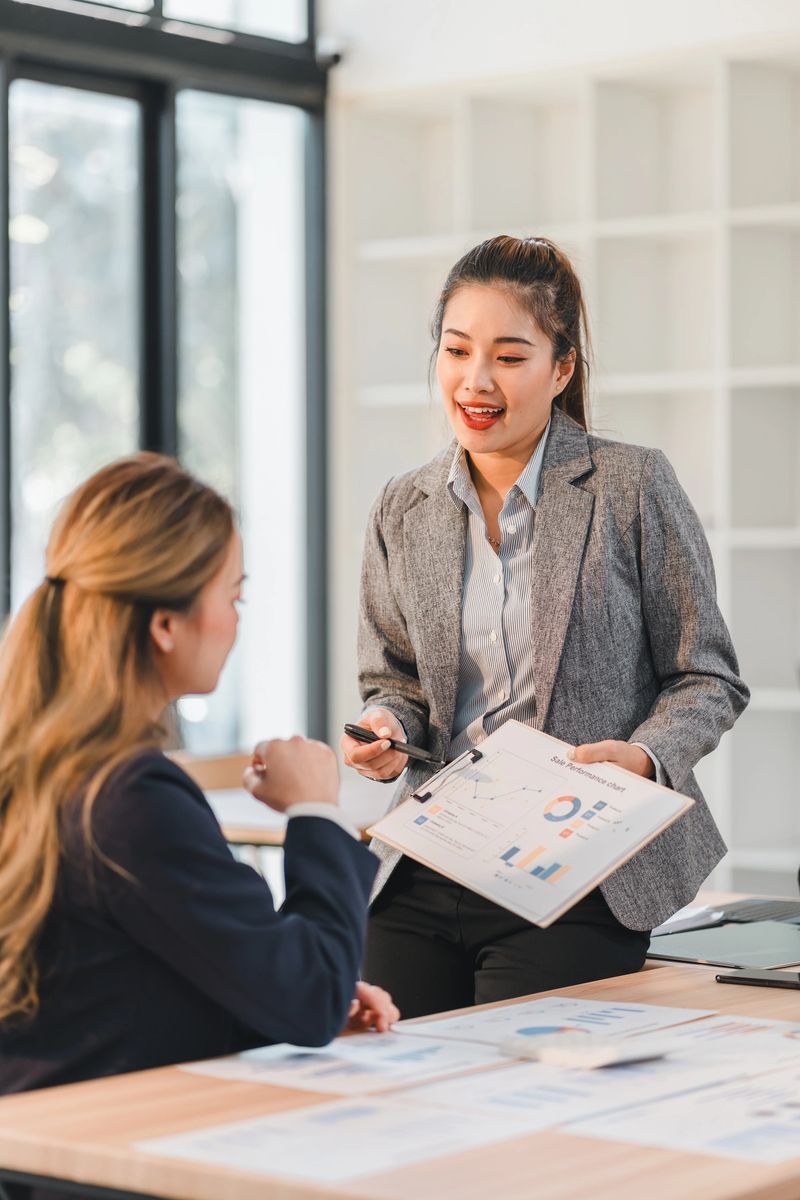 A Two businesswomen discussing financial charts in modern office, with one presenting data on clipboard while other listens attentively. atmosphere is professional and collaborative