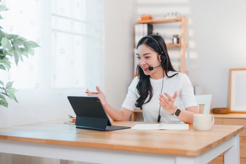 A woman wearing headset conducts video call on tablet at bright home office desk