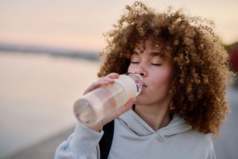 Young curly haired sportswoman drinking water from reusable bottle after exercising outdoors near the river at sunset, enjoying healthy lifestyle