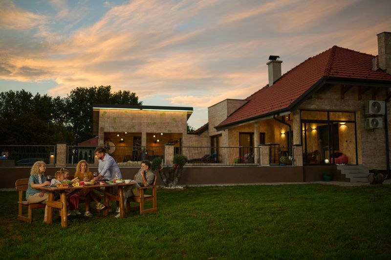 Family is sitting at a wooden table in their backyard, enjoying dinner together as the sun sets. The home is modern and stylish, with a large yard and a warm, inviting atmosphere