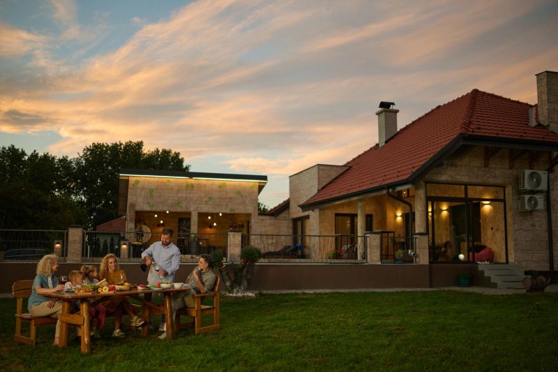 Multigenerational family is enjoying a meal together at an outdoor table in their backyard, with the sun setting behind their home