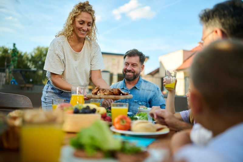 Smiling woman is bringing a platter with grilled chicken to a table full of food, during a sunny family lunch in the backyard