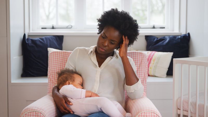 Medium full shot of happy Black mother posing for photo holding little daughter on lap, while sitting with baby girl on couch in cozy childrens bedroom