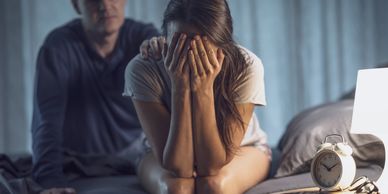 A distressed woman sits on a bed covering her face, with a concerned man behind her.