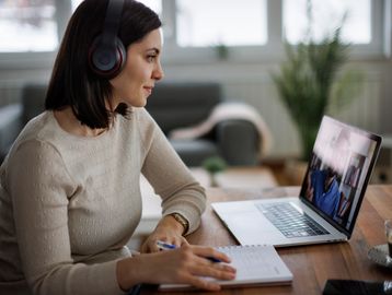 Woman wearing headphones participating in an online video call while taking notes.