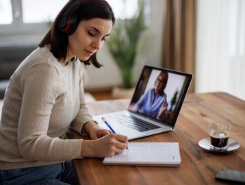 Woman attending an online class, taking notes with headphones on.