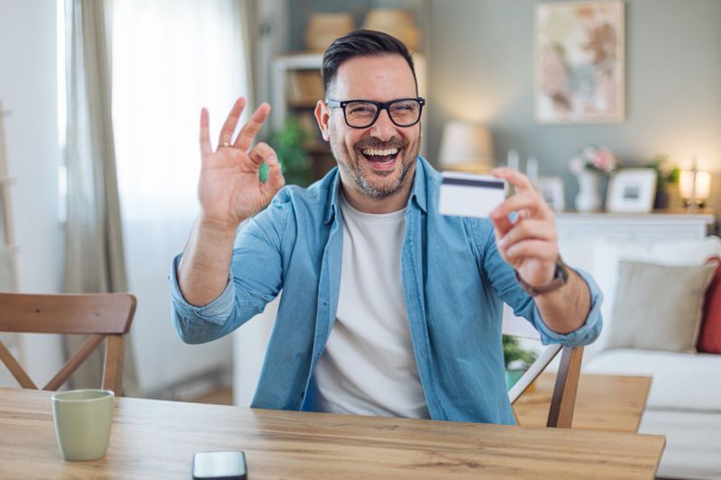 Happy man sitting in the living room, showing OK sign and credit card.