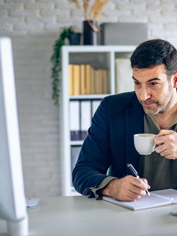 Man drinking coffee while taking notes at his desk.