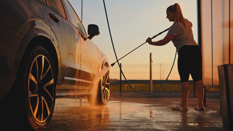 A mid adult Caucasian woman washes a car at a self-service car wash during sunset. She wears casual attire and uses a hose, surrounded by warm sunlight and reflective surfaces.