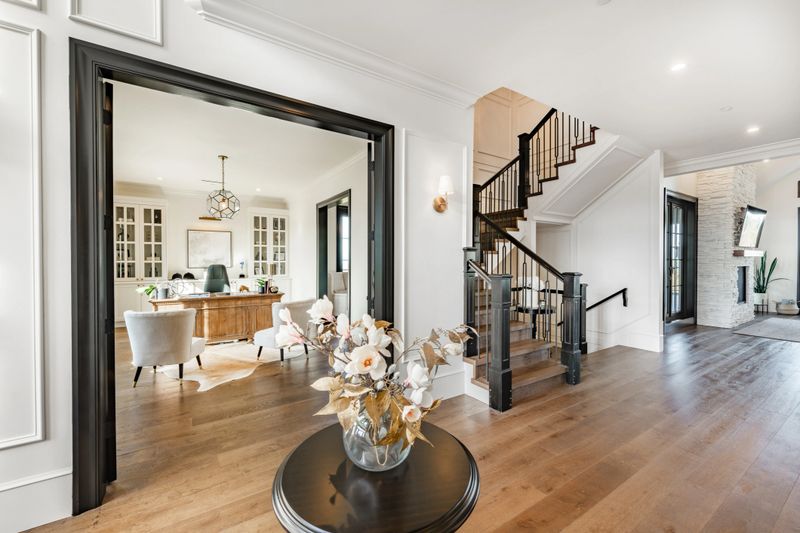 Beautiful large and luxurious estate home interior front door entry hallway foyer with large glass and black wood door expansive entry with a round table containing an arrangement of dried flowers in a bouquet. The view from the front is to a home office and a casual sitting room with blue velvet sofas. large wooden barn doors lead to another room there is a staircase and steps going up.