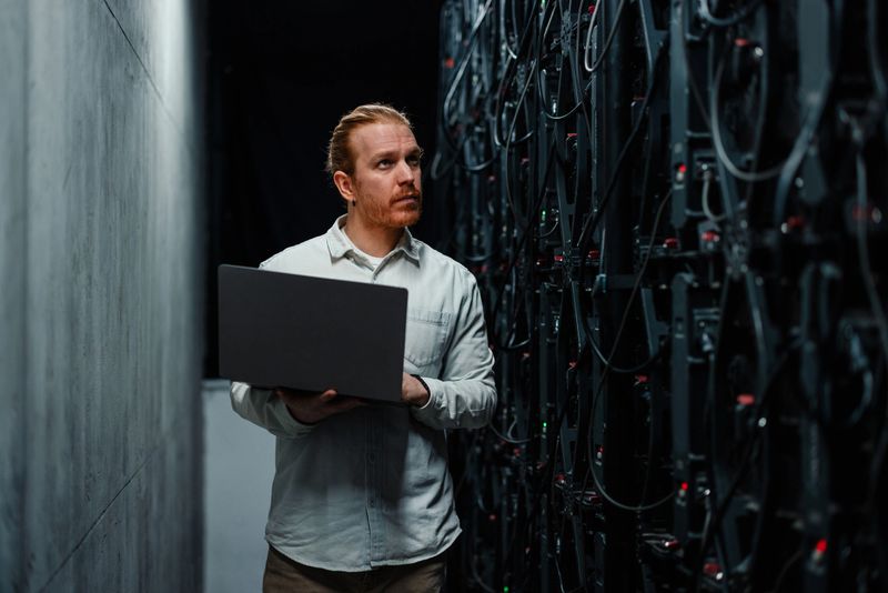 IT specialist working intently on a laptop in a high-tech server room, managing systems and ensuring optimal performance and security