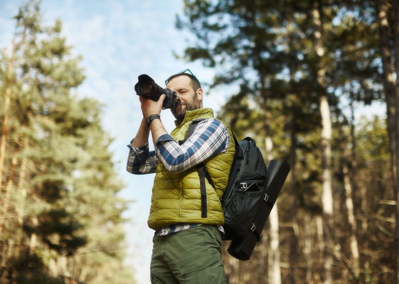 A handsome middle-aged man, a passionate photographer, walks, photographing and explores the splendor of nature in a beautiful pine forest. Looking for a shot for the cover. The man is missing half of the thumb on his right hand. Crouching body position.