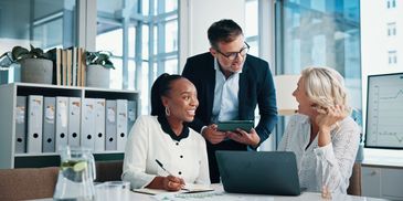 Three colleagues joyfully discuss work around a laptop in a modern office.