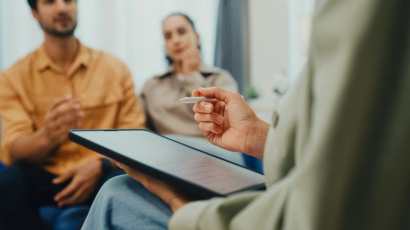 Closeup of young couple in serious discussion with mental health professional during marriage counseling session sit on sofa in living room at home. Therapy session and mental health concept.