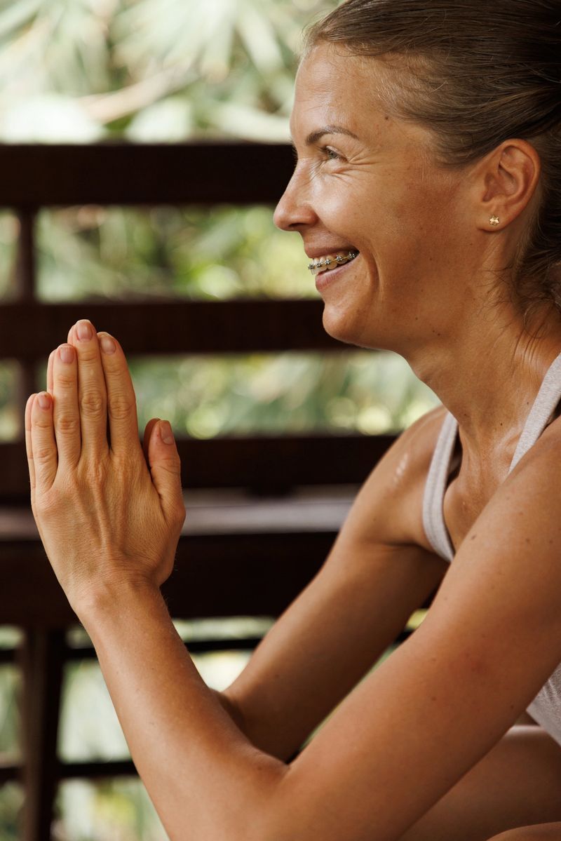 A smiling woman with clasped hands practices mindfulness, emphasizing wellness and gratitude. The peaceful background enhances the theme of calm and focus for meditation or yoga in a serene, natural setting.