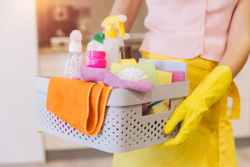 Woman wearing yellow rubber gloves holding basket containing cleaning products and equipment