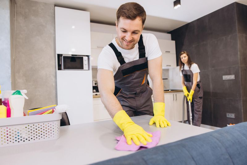 Cleaning company employees wearing uniforms and yellow gloves cleaning a kitchen table and floor