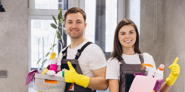 Two cleaners with cleaning supplies and gloves giving thumbs up indoors.
