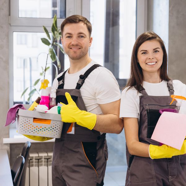Two cleaners in uniforms with cleaning supplies give thumbs up indoors.