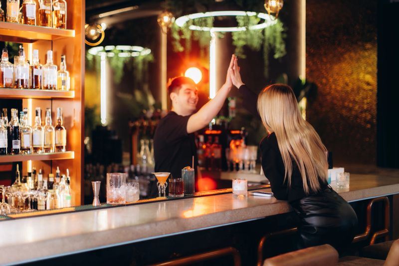Bartender and patron high-fiving in a lively bar setting, surrounded by beverages and neon light decor. A cheerful moment capturing hospitality, interaction, and the festive atmosphere of a trendy lounge.