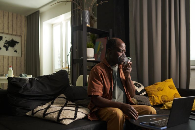 Senior man inhaling fumes from oxygen mask of nebulizer while sitting in front of laptop during online medical consultation