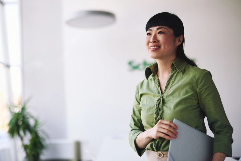 Japanese businesswoman in a green shirt, holding a laptop and smiling while looking away, standing confidently in a modern office filled with natural light and contemporary design elements
