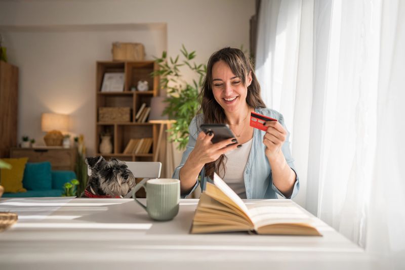 Happy woman shopping online with a smartphone and credit card, sitting comfortably at a table in her cozy living room, enjoying quality time with her dog by her side