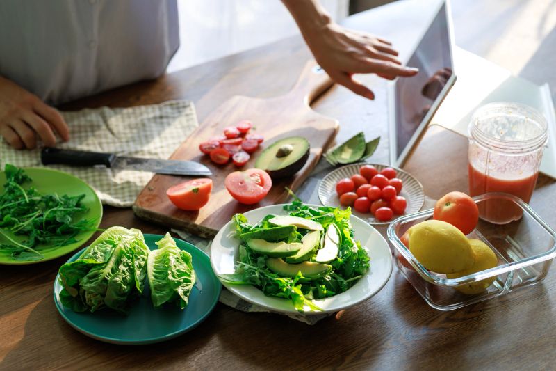 A person preparing a fresh avocado salad with greens, tomatoes, and lemons while using a tablet for guidance. The wooden table is filled with vibrant vegetables, creating a wholesome meal scene.