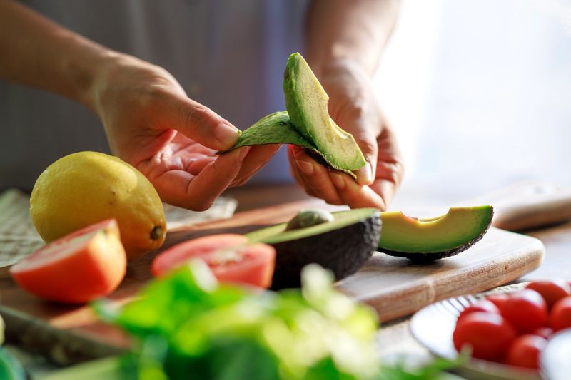 Close-up of hands peeling a ripe avocado over a wooden cutting board, surrounded by fresh ingredients like tomatoes, lemon, and greens. A perfect moment in healthy meal preparation.