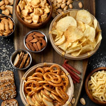 Assorted savory snacks including pretzels, chips, nuts, and crackers arranged on a wooden board.