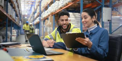 Two warehouse workers discussing work details with a laptop and clipboard.