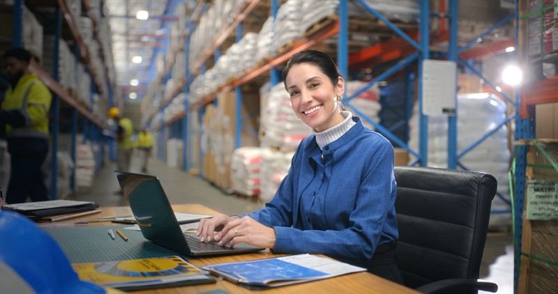 Woman, smile and logistics at warehouse, laptop and typing invoice for delivery, stock and inventory. Person, portrait and computer for supply chain management, cargo or distribution at storage plant