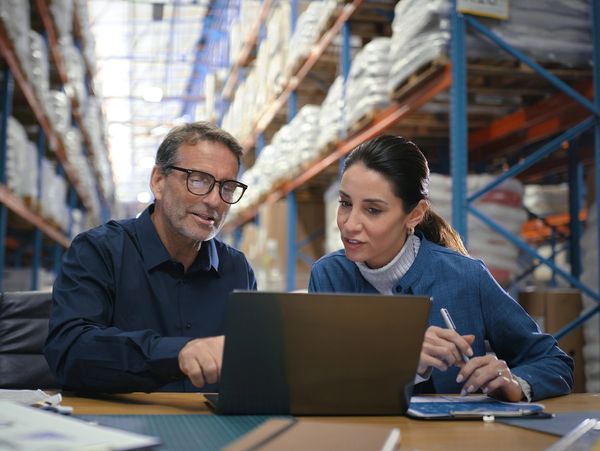 Two colleagues review a laptop in a warehouse office.